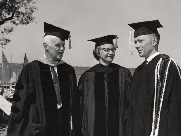 Three people, two men and one woman, talk outdoors while dressed in academic regalia.