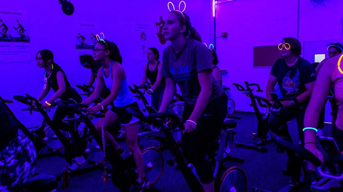 Students riding indoor bicycles in a fitness room with purple-hued lighting effects. The students are wearing a variety of glow-in-the-dark accessories on the heads and wrists.