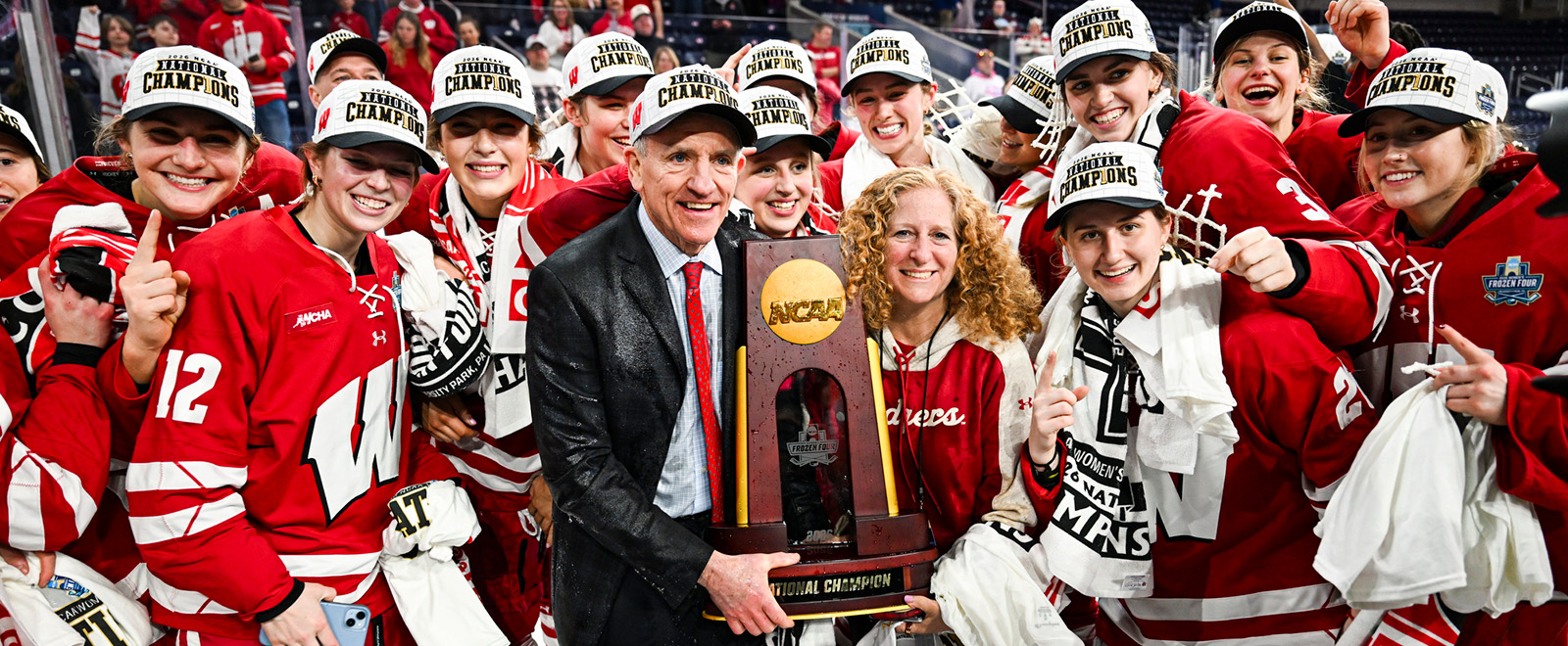 Coach Mark Johnson and Chancellor Jennifer Mnookin both hold the Women's Hockey NCAA championship trophy and smile surrounded by the UW Women's Badger hockey team