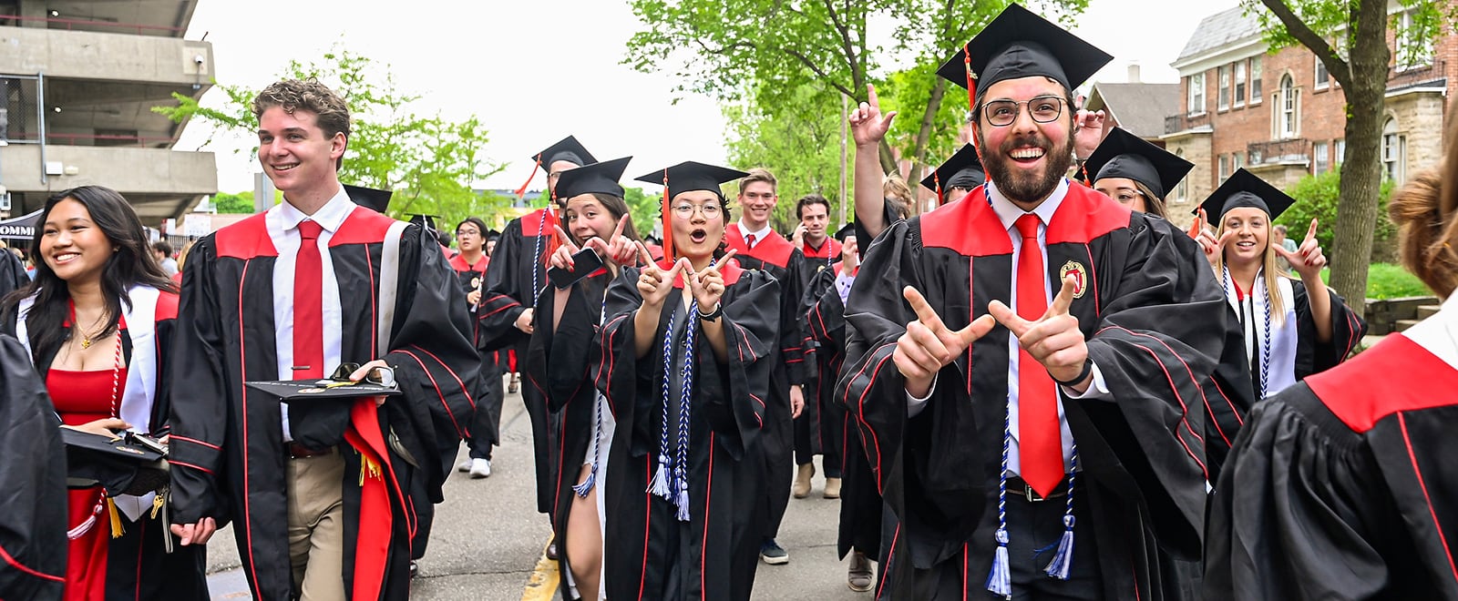 A group of happy graduates wearing black caps and gowns with red stoles walk outdoors along a campus street, some holding mortarboards and making a 'W' hand gesture.