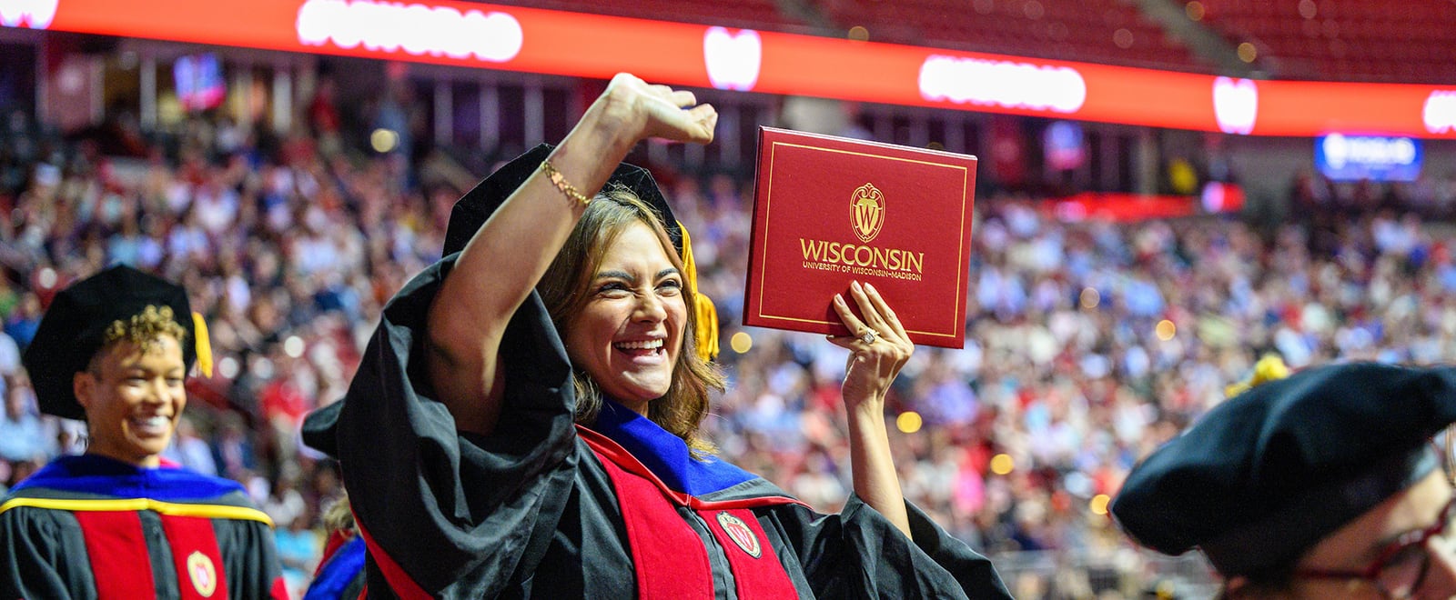 A graduate wearing academic regalia holds a red diploma cover reading 'Wisconsin' above shoulder height as other graduates and a large audience fill the the Kohl Center in the background.