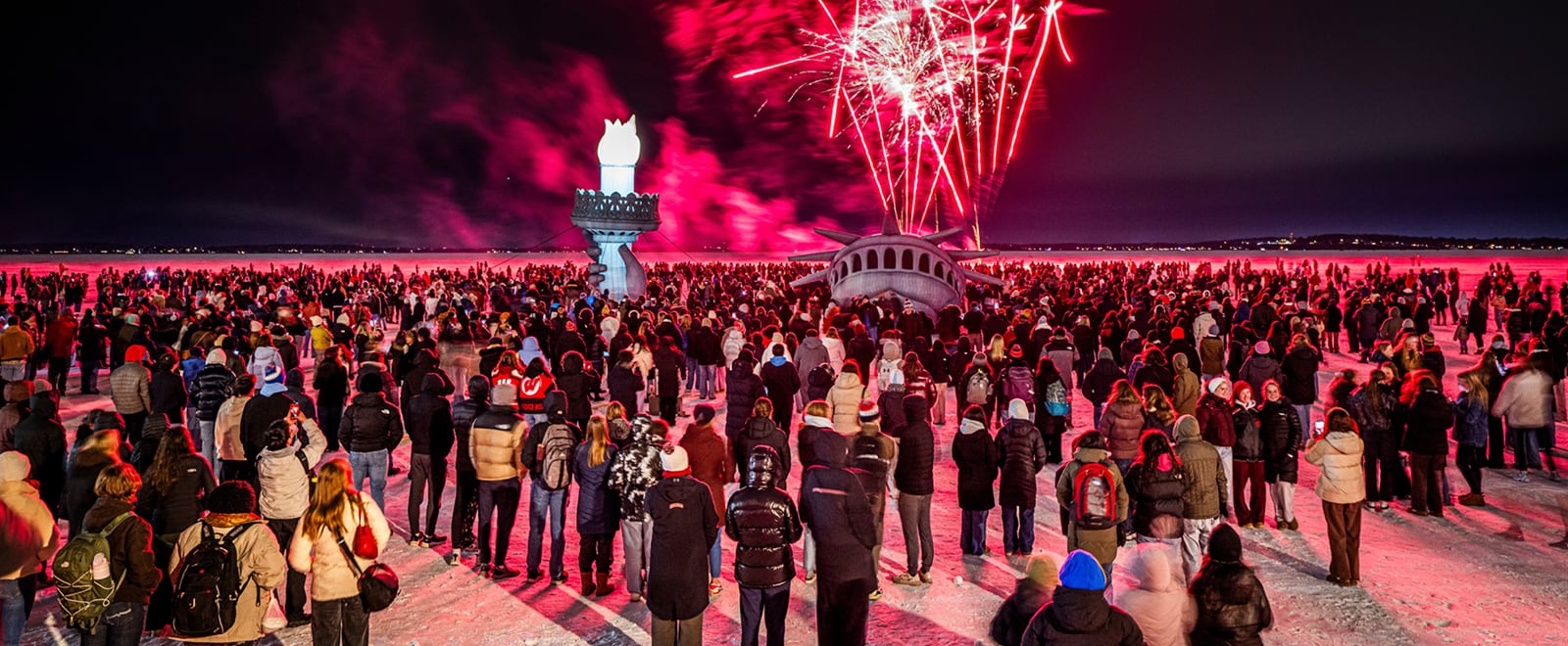 A large crowd gathered outdoors at night around the Lady of the Lake statue on frozen Lake Mendota to watch bright red fireworks bursting in the sky.