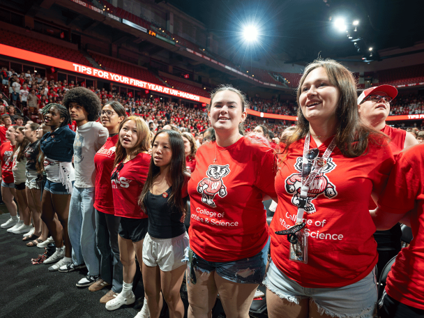 A line of smiling students wearing Badger gear stand side-by-side on an arena floor during a UW-Madison orientation event.