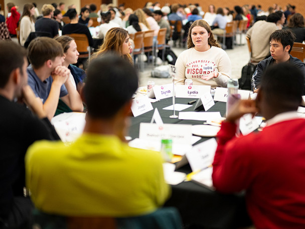 Numerous groups of people sitting at round tables with name cards, in conversation.
