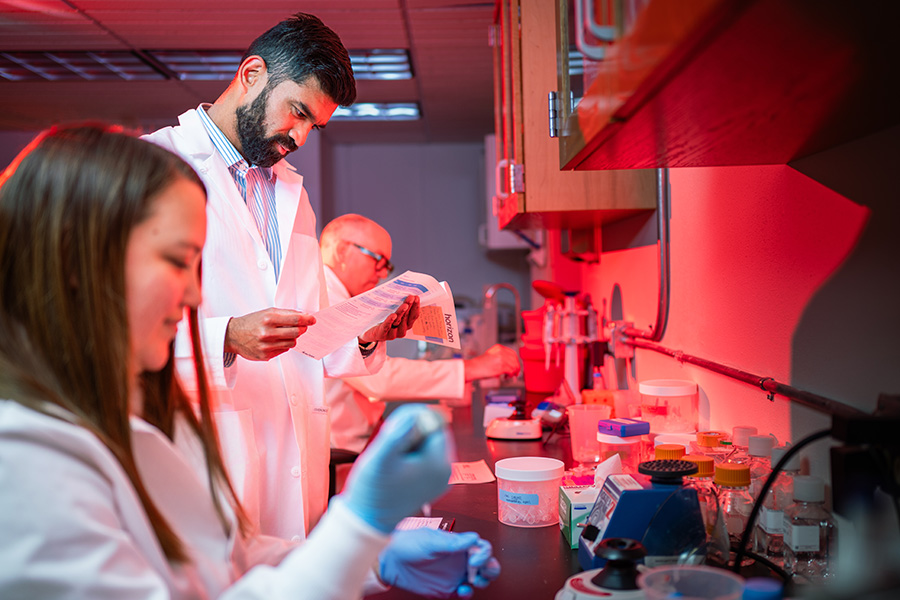 In a red-lit laboratory, three people in white lab coats conduct research -- one reads a document, another uses a pipette, and the lab counter is filled with scientific equipment.