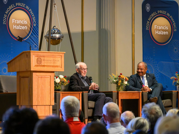 Two people sitting on a stage with microphones speaking to a crowd
