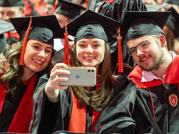 Three graduates in academic regalia lean together to take a selfie.
