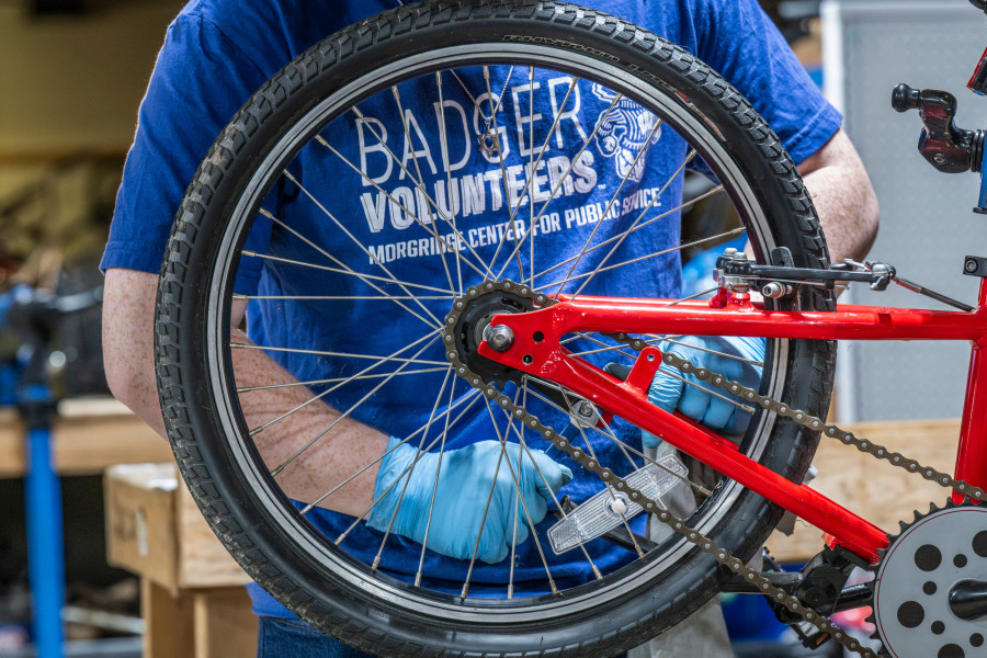 A person wearing blue gloves adjusts the rear wheel and chain of a red bicycle, with a blue shirt reading “Badger Volunteers, Morgridge Center for Public Service” visible in a workshop setting.
