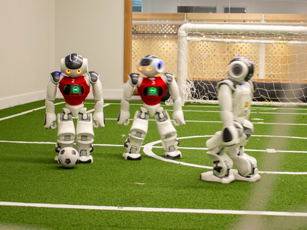 Three small humanoid robots play soccer on a miniature green turf field with white markings and a small goal in the background.