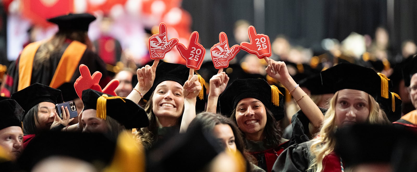 Graduates in black caps and gowns at a commencement ceremony holding up red foam fingers -- one has an image of Bucky Badger, and the other reads ‘2025’.