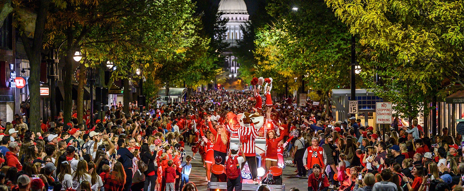 Bucky Badger and the Spirit Squad rally the crowd as they travel down State Street during the annual evening Homecoming Parade, with the lighted state capitol building in the background.