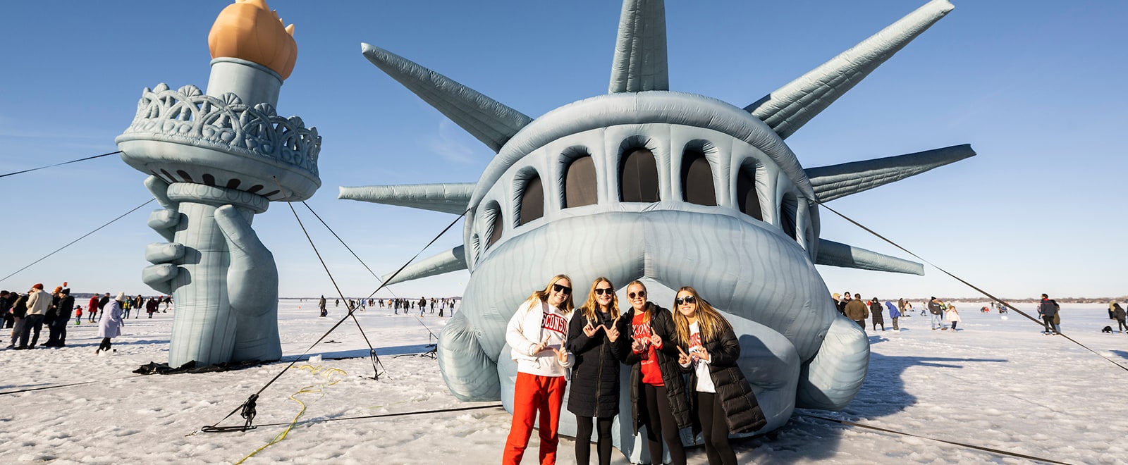 Four students stand on frozen Lake Mendota and flash the 'W' sign in front of the 'Lady of the Lake' -- what appears to be a mostly-sunken Statue of Liberty.