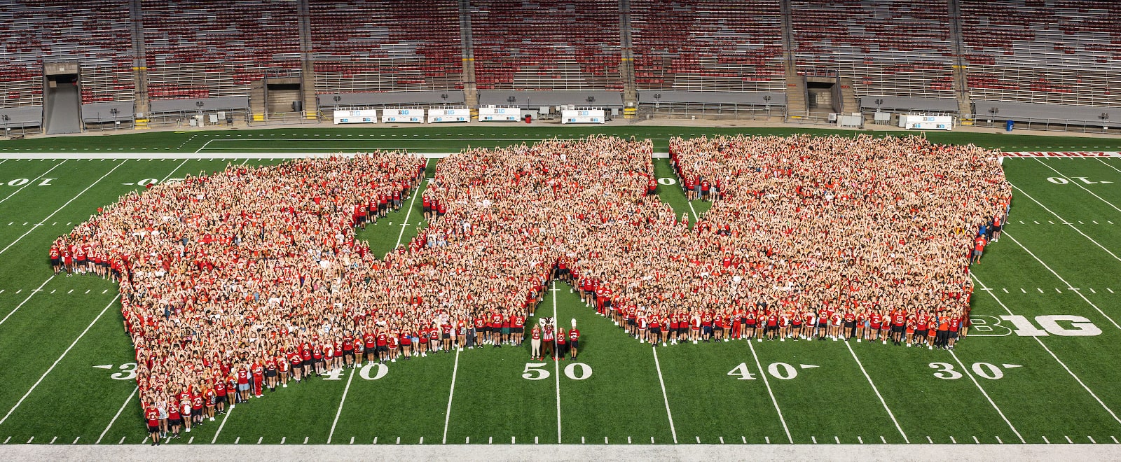 Thousands of UW students stand on the football field at Camp Randall Stadium, forming the shape of UW Athletic's Motion W logo.