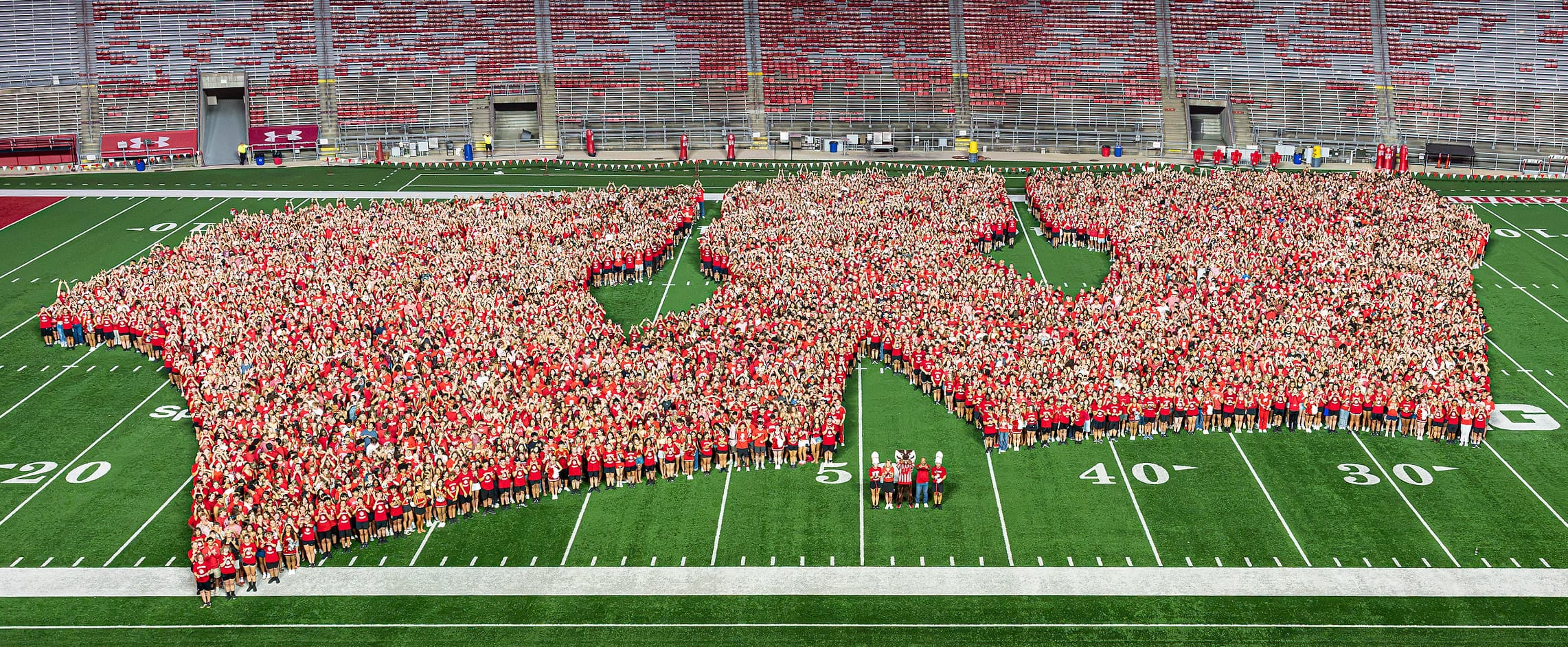 An overhead wide shot of thousands of incoming students wearing red shirts assembled on the field at Camp Randall Stadium to form a massive Motion W logo.