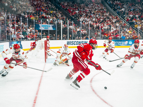 Six Denver players wearing white try to defend their goal against one Wisconsin player in red. Wisconsin's player has the pick and is looking to pass to a teammate.