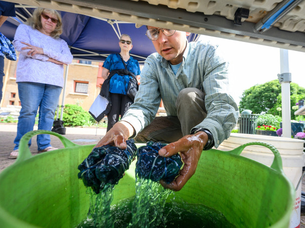 A man pulls two pieces of tie-dyed clothing from a large bucket of water while people look on behind him.