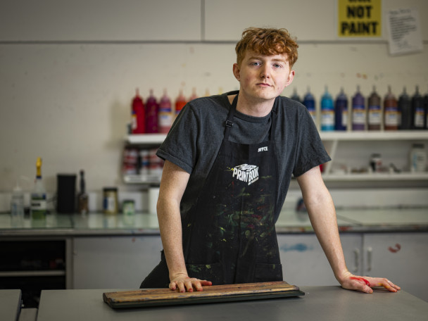 Photo of Bryce Dailey in a workshop apron in a screen printing studio as he leans with his hands on a work table.