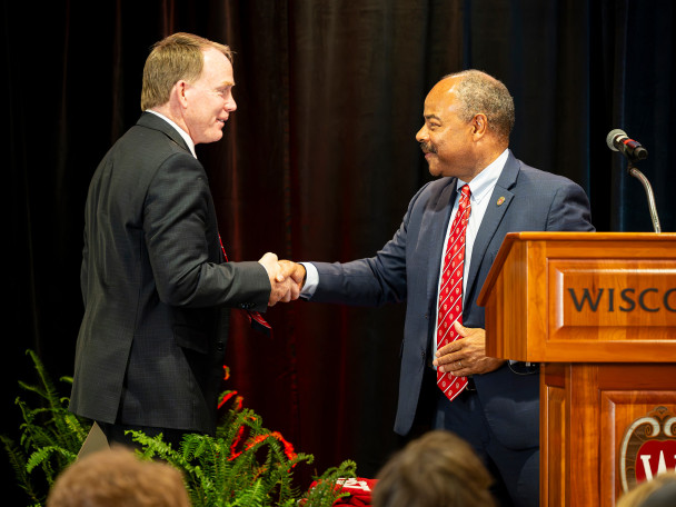 Shawn Eichorst stands on the left and shakes hands with interim Chancellor Eric Wilcots, who stands on the right behind a wooden University of Wisconsin podium.