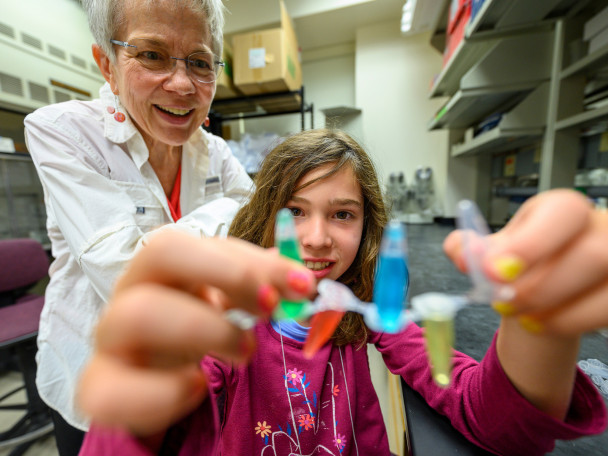 A young girl holds up a strip of plastic tubes containing colorful liquids while a woman with short grey hair watches and smiles in a laboratory setting.