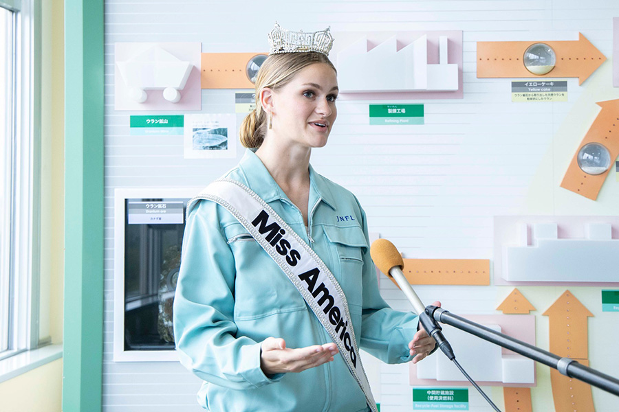 A woman in a jumpsuit wearing a tiara and a sash that says Miss America stands and speaks at a microphone.