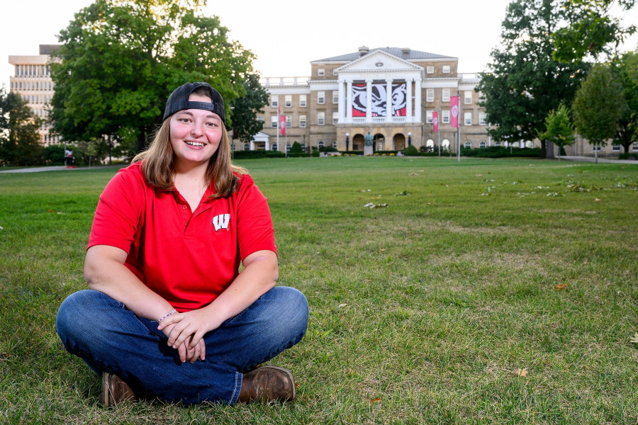 A woman in a red shirt with a "W" logo and jeans sits on the spacious lawn in front of Bascom Hall.