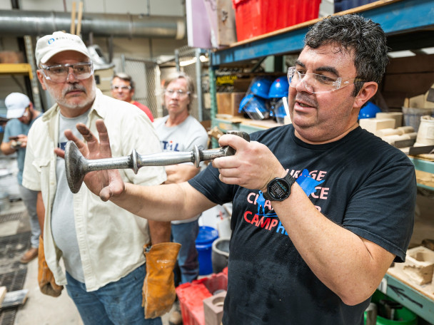 Herrera wears safety glasses and holds a molded piece of aluminum while talking bootcamp participants.