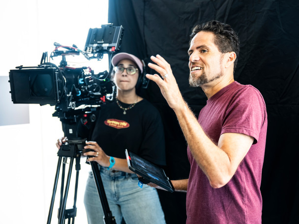 A filmmaker is talking with a digital tablet in hand, while a person operating a video camera looks on