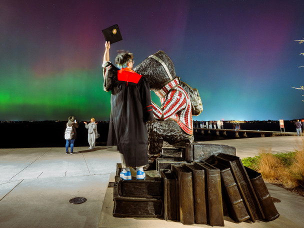 A student wearing a graduation gown stands next to a Bucky Badger statue while holding his cap to the sky. In front of him, Northern Lights dance across the sky.