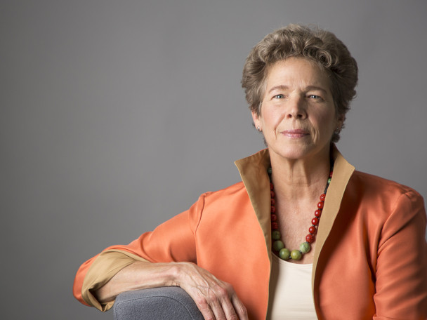 Portrait photo of Tia Nelson against a gray backround. She is wearing an orange blazer with a high collar and a green and red beaded necklace