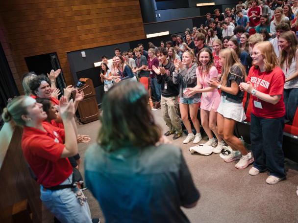A row of UW-Madison students, all wearing bright Badger red shirts, stand in front of a full auditorium and lead the room of new students in a clapping chant.