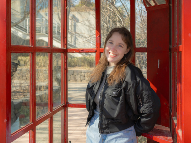 A woman is shown in a telephone booth painted red, with a building visible in the background.