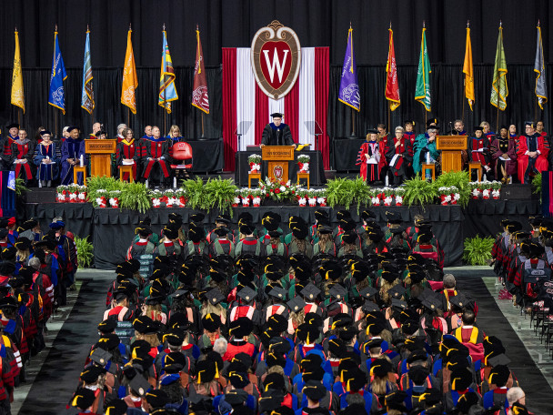 Shown from the crowd’s perspective, Provost John Zumbrunnen speaks at the podium on the commencement stage, flanked by seated members of the official party.
