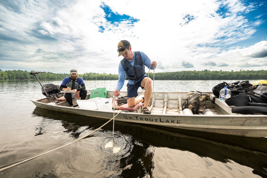 Jack Abel kneels at the side of a low fishing boat to lower a rope into the water. His research partner, Quinn Smith, sits in the back of the boat and takes notes on a clipboard.