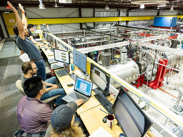 A half-dozen people sit at a control panel area overlooking equipment. One of them stands and raises his hands in celebration.