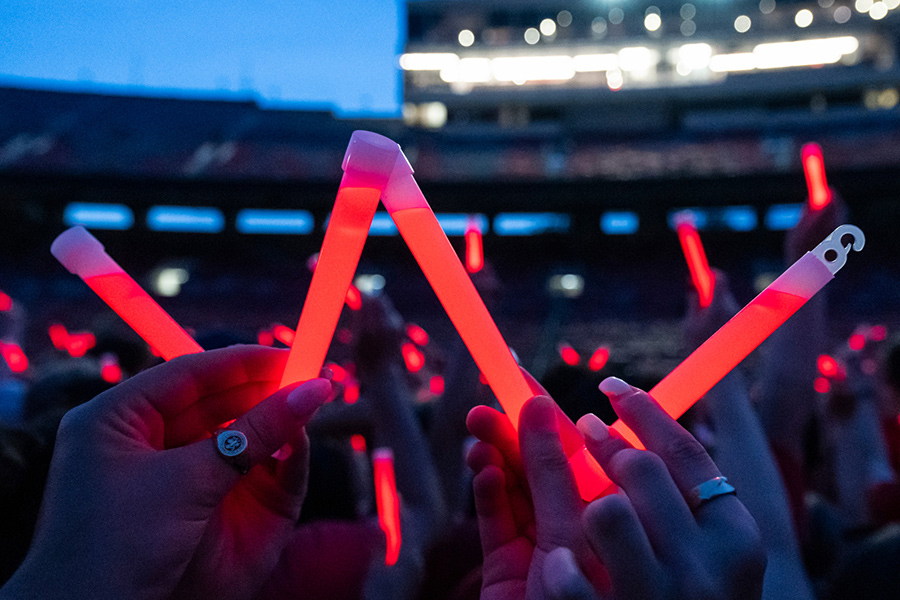 Audience members holding up glowing red sticks at a nighttime event in Camp Randall Stadium. In the foreground, someone holds the sticks to form a letter W.