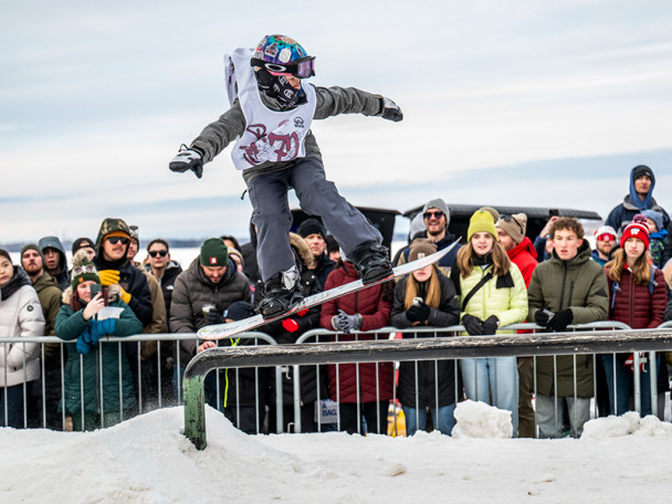 A snowboarder performs a rail slide as a bundled‑up crowd watches from behind a barricade.