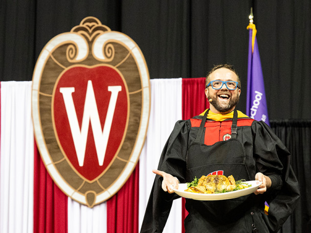 Photo of Dan Jacobs in academic robes holding up a roast chicken on the commencement stage.