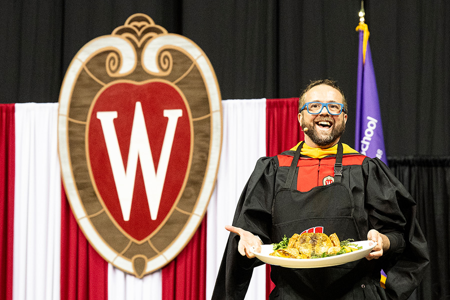 Photo of Dan Jacobs in academic robes holding up a roast chicken on the commencement stage.