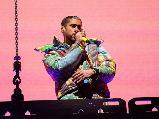 Bad Bunny, wearing a colorful jacket against a bright pink background, performs with a microphone in hand.