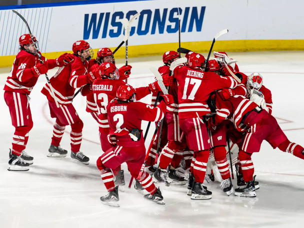 A group of Wisconsin Badgers men's hockey players in red and white uniforms celebrate a victory by huddling together and cheering on the ice.