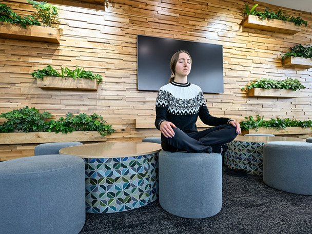 A woman sits cross-legged with her eyes closed on a round, gray ottoman in a modern lounge area with several planters mounted on the wall behind her.