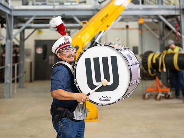 A man wearing a UW Marching band cap beats a band-branded bass drum in the unlikely setting of a steamfitters training facility.