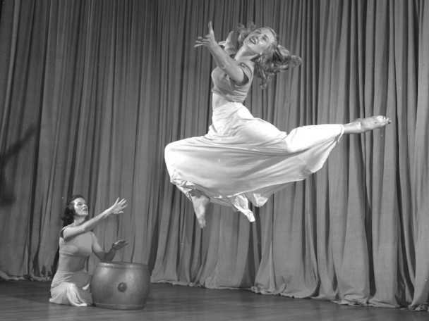 Grayscale photo of a dancer leaping gracefully across a stage in flowing attire while another performer sits nearby playing a drum.