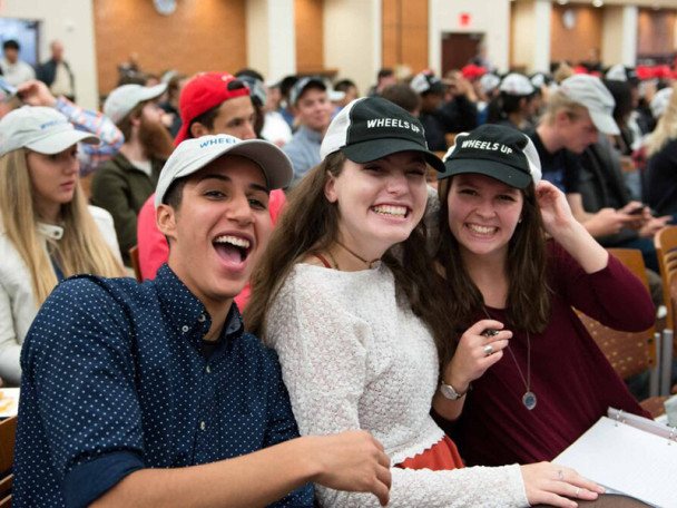 Three seated students wearing ball caps with the words 'Wheels Up' are in the foreground of a lecture hall filled with seated students.