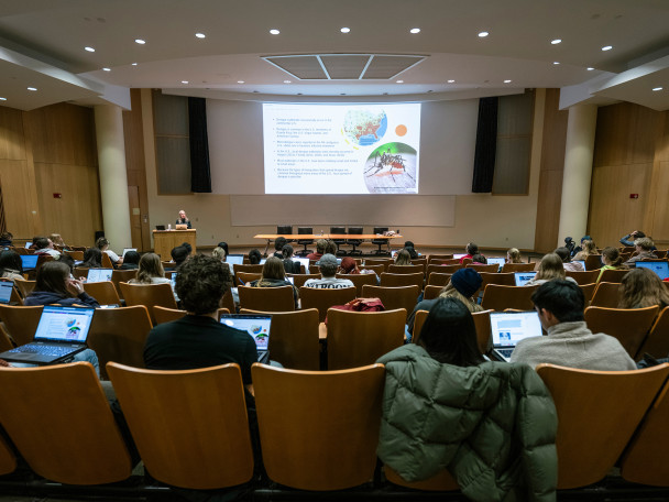 A photo take from the back of an auditorium-style classroom shoes students watching a power point slide while a professor talks at the front of the room.