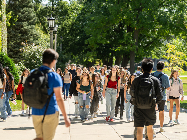 Several young men and women carrying backpacks walk up a tree-lined sidewalk.