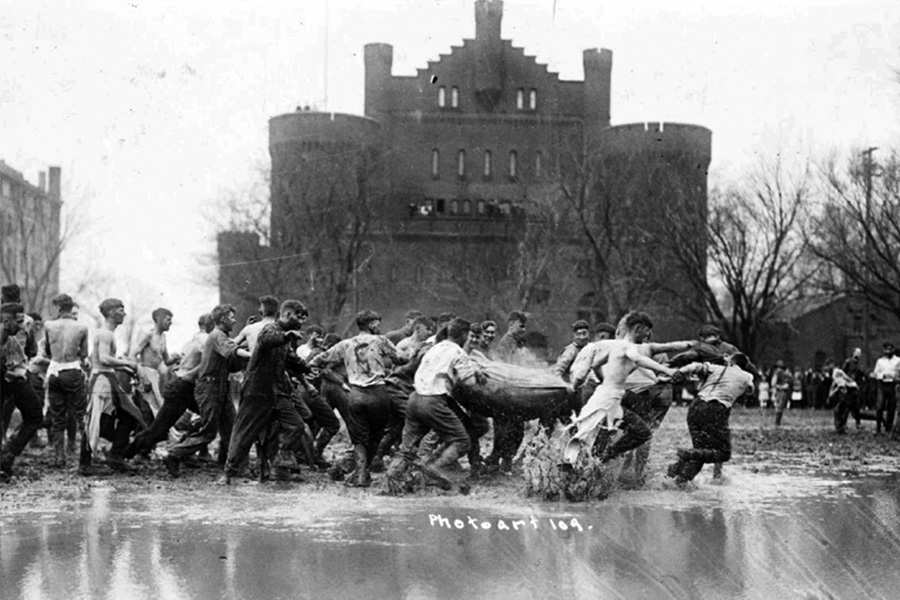 A historic black‑and‑white photograph of a group of young men engaged in a muddy game with a large sack in rain‑soaked field Library Mall. The Red Gym is in the background.