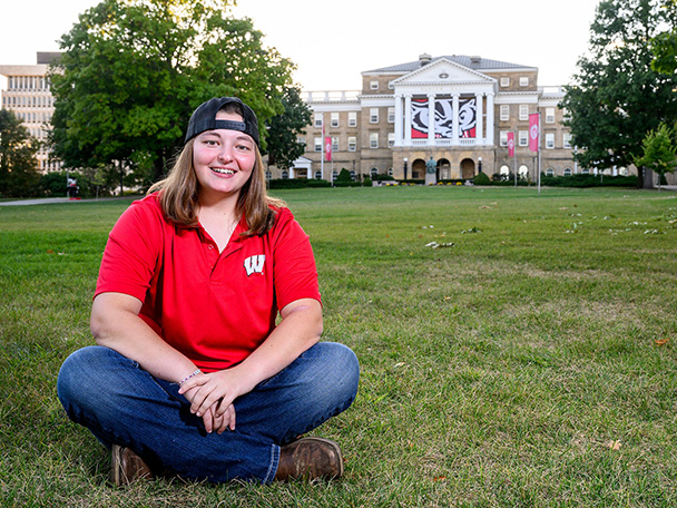 A woman in a red shirt with a "W" logo and jeans sits on the spacious lawn in front of Bascom Hall.