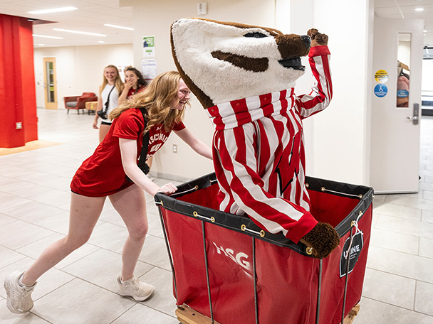 UW-Madison mascot Bucky Badger rides in a big red cart inside of a residence hall. He is being pushed by a student wearing a red Wisconsin Badgers t-shirt.