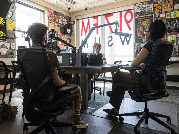 Three students sit in front of microphones in a radio station.